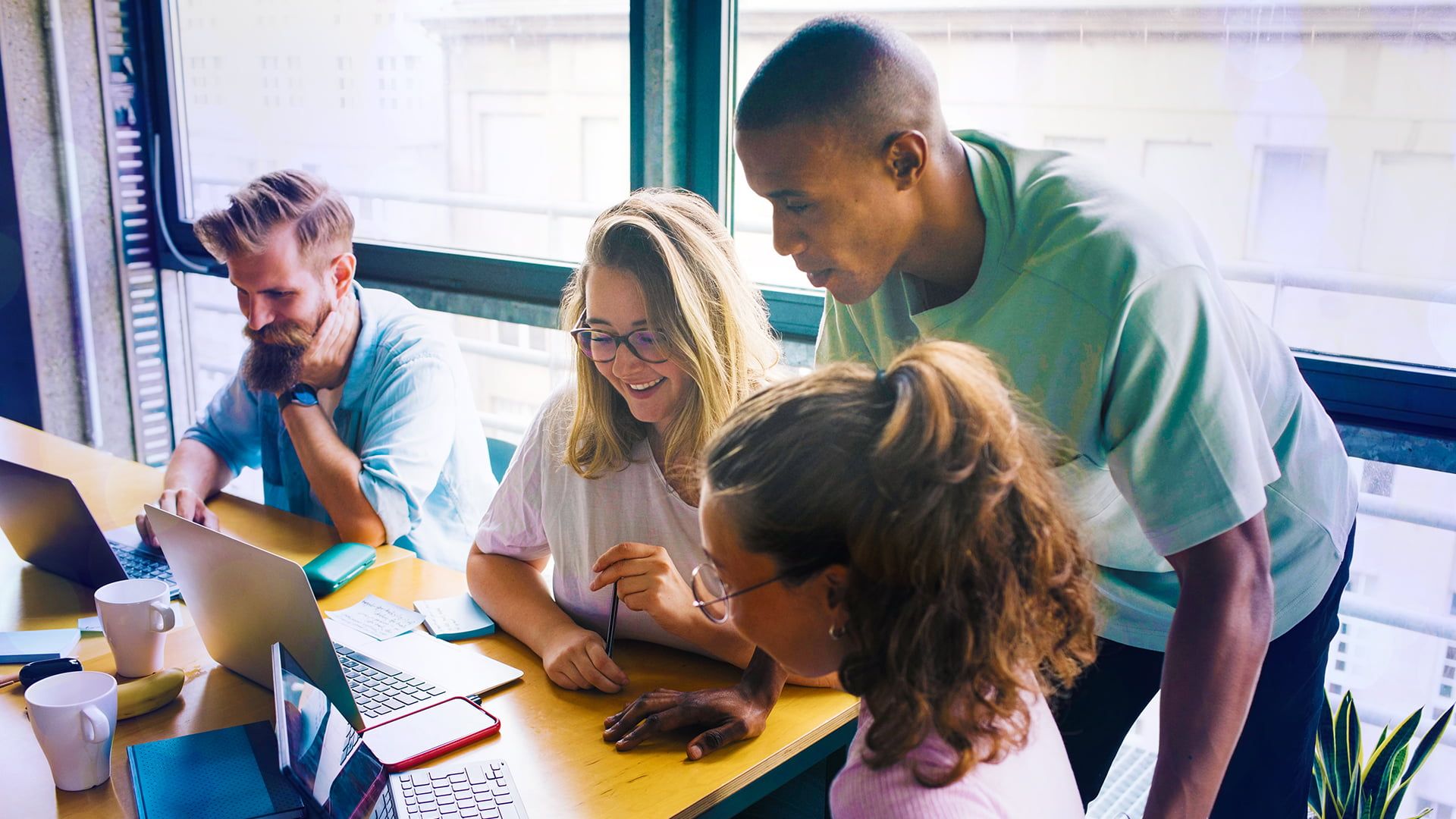 En grupp på fyra personer som samarbetar runt ett bord, använder bärbara datorer och ler, med ett fönster och utsikt över staden i bakgrunden.